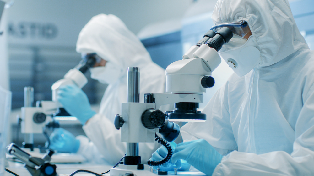 2 scientists in protective gear inspecting particles under a microscope.